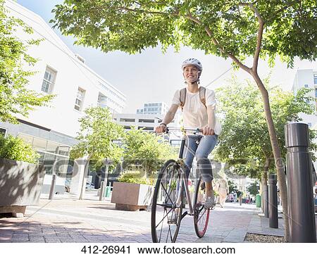 Stock Image - Smiling young woman with helmet riding bicycle in urban park. Fotosearch