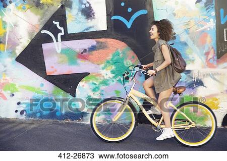 Stock Image - Woman riding bicycle along urban multicolor graffiti wall. Fotosearch
