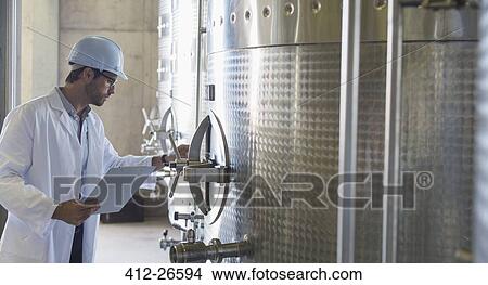 Picture - Vintner in lab coat and hard hat examining stainless steel vat in cellar. Fotosearch