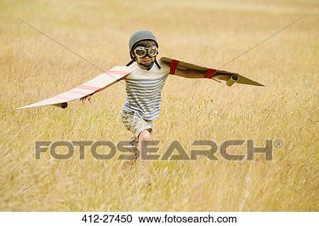 Stock Image - Boy running with wings and aviator’s cap and flying goggles in field. Fotosearch