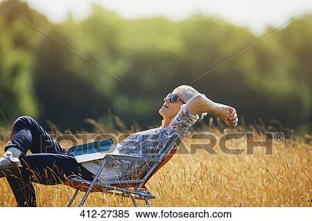 Stock Photography - Carefree senior woman relaxing with book in sunny field. Fotosearch