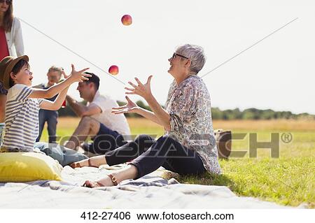 Stock Photograph - Grandmother and grandson juggling apples on picnic blanket in sunny field. Fotosearch