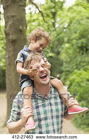 Stock Image - Playful son covering father’s eyes in woods. Fotosearch