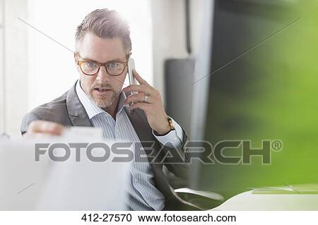 Businessman examining paperwork talking on cell phone in office View Large Photo Image Stock Image - Businessman examining paperwork talking on cell phone in office. Fotosearch
