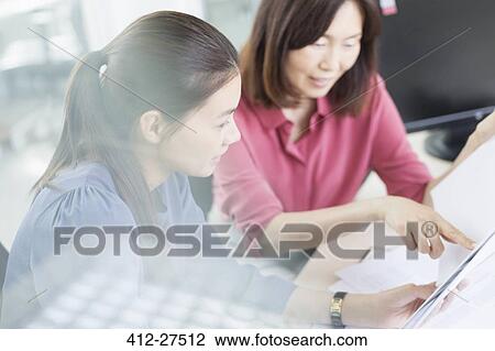 Stock Image - Businesswomen discussing paperwork in office. Fotosearch