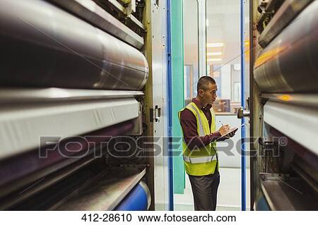 Stock Image - Engineer with clipboard examining printing press machinery. Fotosearch