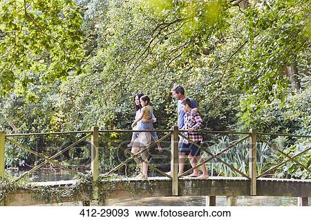 Stock Image - Family crossing footbridge in park with trees. Fotosearch