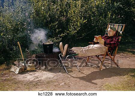 Stock Photo - 1950S 1959 Man Reading Sitting Folding Camp Chair Campfire Grill Pot Smoke Axe Fire Wood Pattison State Park Wisconsin. Fotosearch
