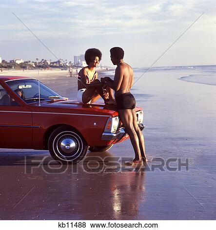 Stock Photo - 1970 1970S African-American Couple Beach Woman Sitting Hood Car Man Holding Her Hands Romance Vacation Honeymoon Romance . Fotosearch