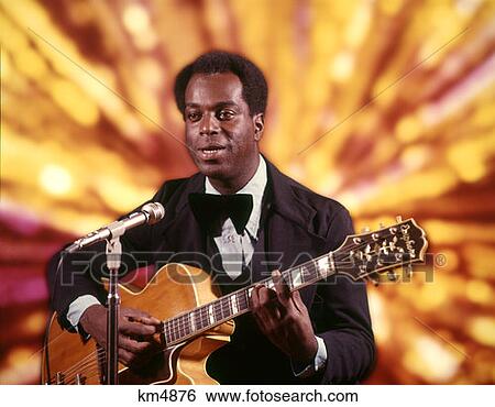 Stock Photograph - 1970 1970S African-American Man Playing Guitar Singing Microphone Tuxedo Gold Lights Background Singer Entertainer . Fotosearch