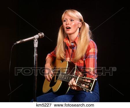 Stock Photo - 1970 1970S Blond Blonde Young Woman Pigtails Plaid Shirt Jeans Play Guitar Singing Microphone Folk Singer Singers . Fotosearch