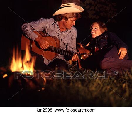 Stock Photo - 1970 1970S Couple Campfire Night Man Wearing Cowboy Hat Playing Guitar Music Country Western Woman Men Women Couples . Fotosearch