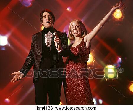 Stock Image - 1970 1970S Duo Man Woman Couple Singing Woman Red Dress Gown Man Tuxedo Holding Microphone Stage Lights Background . Fotosearch