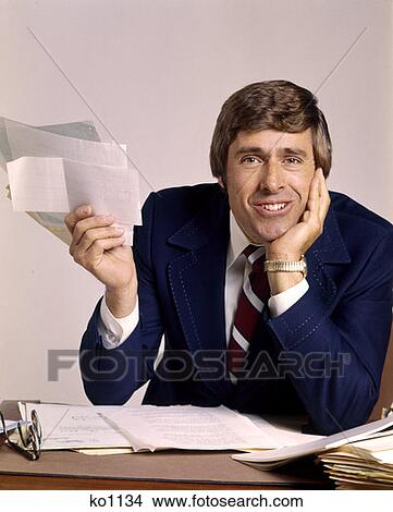 1970 1970S Man Businessman Business Desk Holding Papers Paperwork Bills Smiling Funny Face Expression View Large Photo Image Picture - 1970 1970S Man Businessman Business Desk Holding Papers Paperwork Bills Smiling Funny Face Expression . Fotosearch