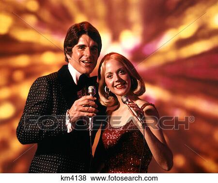 Stock Photo - 1970 1970S Man Woman Singers Singing Vocalist Woman Blonde Red Dress Hold Microphone Man Tuxedo Entertainers . Fotosearch