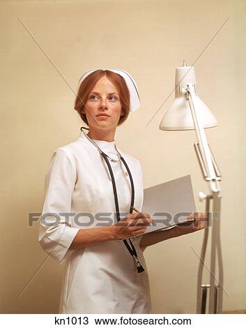 Stock Image - 1970 1970S Nurse Holding Clipboard Wearing Stethoscope White Uniform Cap Lamp Nurses Medical Medicine Career . Fotosearch