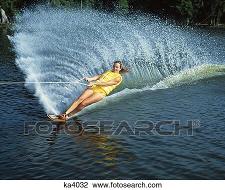 Stock Image - 1970 1970S Young Woman Water Skiing Trhowing Up Water Fan Splash Wearing Life Jacket Yellow Swimsuit Sports. Fotosearch