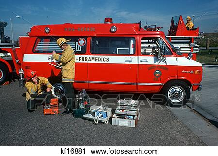 Stock Image - 1970S 1980S Two Men Paramedic Team Inspecting Checking Equipment Next To Ambulance Van Wearing Fireman Helmets. Fotosearch