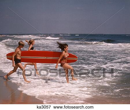 Stock Photo - 1970S 3 Teen Boy Two Girls In Bathing Suits Running Into Surf Carrying A Red Surfboard Laughing Beach Sand Ocean Fun . Fotosearch