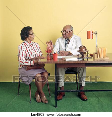 Stock Image - 1970S African-American Man Doctor Talking With An African-American Woman Patient At Desk. Fotosearch