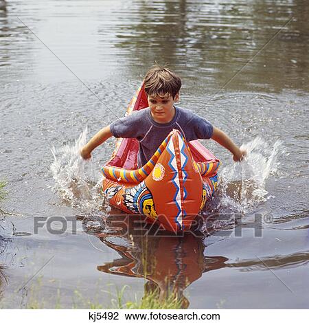 Stock Image - 1970S Brunette Boy In Blue Shirt In Red Inflatable Canoe Indian Motif Paddle Splashing With His Hands Fun Lake Pond. Fotosearch