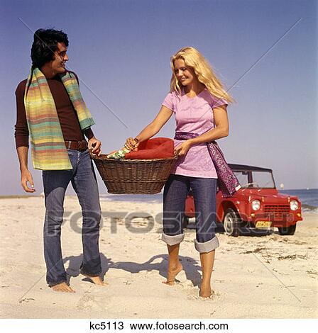 Stock Image - 1970S Couple Beach Carry Picnic Basket Red Dune Buggy In Background. Fotosearch