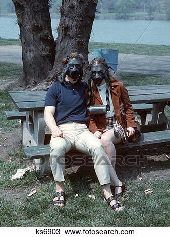Stock Image - 1970S Couple Sitting On Park Bench Wearing Gas Masks. Fotosearch