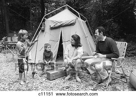 Stock Image - 1970S Family Of Four Sitting In Front Of Tent In Woods Son Holding Fishing Pole In One Hand & Big Fish In Other . Fotosearch