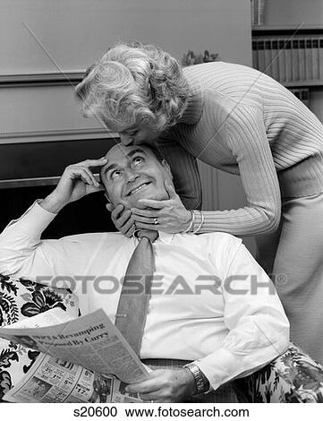 1970S Older Couple Wife Leans Over To Kiss Husband On Forehead He Is Sitting In Chair With Newspaper Indoor View Large Photo Image Stock Image - 1970S Older Couple Wife Leans Over To Kiss Husband On Forehead He Is Sitting In Chair With Newspaper Indoor . Fotosearch