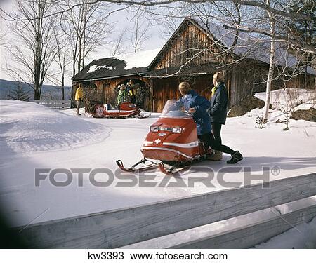 Stock Image - 1970S People In Skimobile Snow Vehicle Outside Rural Cabin Several Woman Man Vacation Fun Lifestyle. Fotosearch
