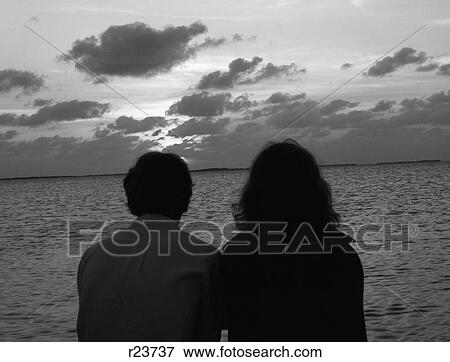 Stock Photo - 1970S Rear View Silhouette Of Man & Woman Sitting At Side Of Water Watching Sunset . Fotosearch