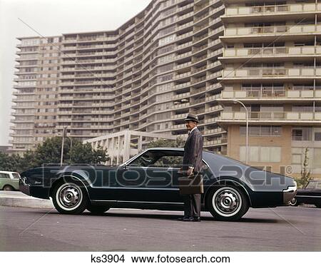 Picture - 1970S Salesman With Attache Case Standing Beside Car Outside Apartment Condominium Building . Fotosearch
