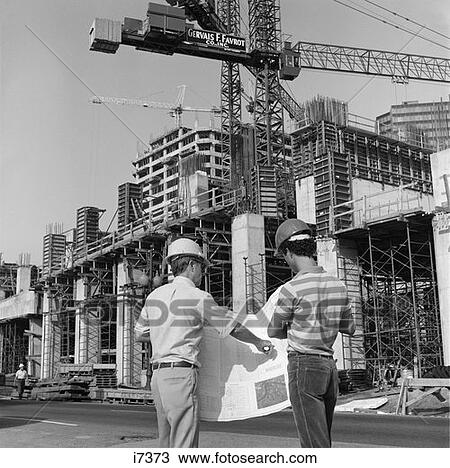 Stock Image - 1980S Rear View Of Two Hard Hat Workers Standing In Front Of Major Construction Site Looking Over Building Plans Men Outdoor. Fotosearch