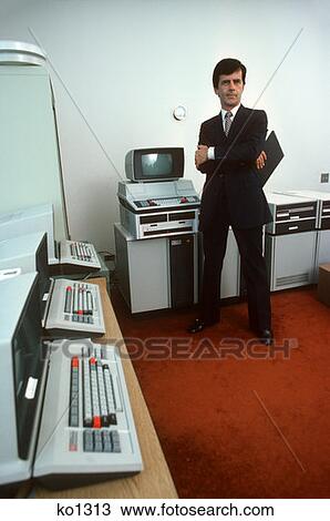 Stock Image - 1980S Retro Man Computer Programmer Standing In A Room With Early Texas Instruments Hardware Screens Keyboards. Fotosearch