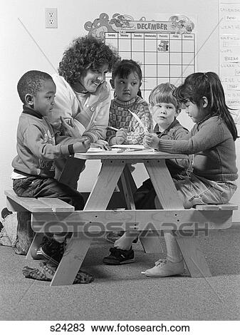 Stock Image - Boys Girls Students Elementary School Sit At Picnic Bench Table Teacher Woman Kindergarten Toddlers Day Care Nursery School. Fotosearch