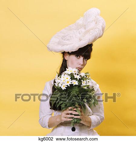 Stock Image - Brunette Woman In 19Th Century Costume Large Silly White Hat Holding A Bouquet Of Daisies Innocent Victorian Flirt. Fotosearch