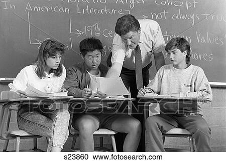 Stock Image - College Students Men Woman Teacher In Classroom Sitting At Desks Teacher Leaning Over Desk Examining Paper Of Student Chalkboard. Fotosearch