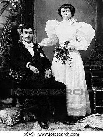 Stock Image - Early 1900S Formal Portrait Of Bride And Groom Man Seated Woman Standing Indoor . Fotosearch