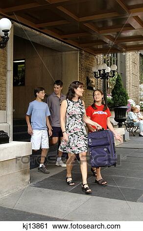 Stock Image - Family Of Four Leaving Hotel Carrying Luggage, Tourists. Fotosearch