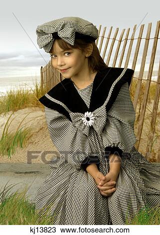 Stock Image - Girl Wearing Old Fashioned Black Gingham Dress And Cap Sitting By Fence On Beach Sand Dune . Fotosearch
