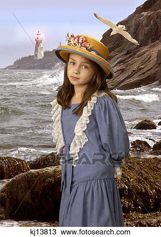 Stock Image - Girl Wearing Old Fashioned Blue Dress And Straw Hat With Flowers Standing By Rocky Beach Shore . Fotosearch