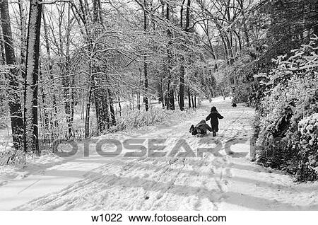 Stock Image - Two Children Walking Down Winter Snow Covered Lane Road Rural 1 Child Walking Other On Sled Footprints . Fotosearch