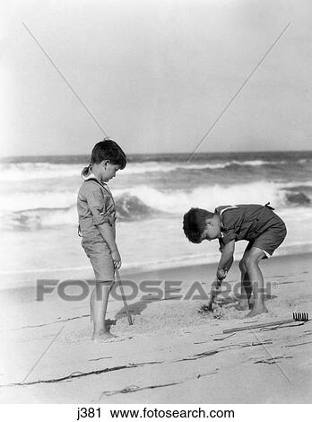Stock Image - 1920 1920S Two Boys With Sand Rake Tools Sticks One Boy Digging In Sand Waves Ocean Surf Shore Summer Fun Playing Retro. Fotosearch
