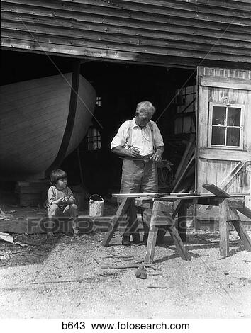 Stock Image - 1920S 1930S Elderly Man Grandfather Working At Saw Horses As Little Boy Watches Hull Of Boat Visible In Background. Fotosearch
