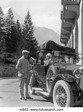 Stock Image - 1920S Couple Man Standing Beside Car Woman Driving Roadster Touring Car Top Full Of Covered Luggage . Fotosearch