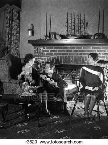 Stock Image - 1920S Man Sitting Between Two Women Flirting With One Woman Ignoring The Other Drinking Tea Fireplace In Background. Fotosearch