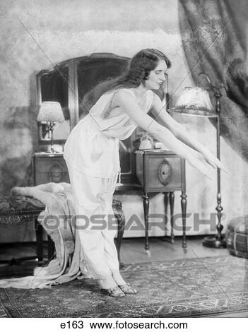 Stock Image - 1920S Woman Exercising In Bedroom Wear Pajamas Arms Stretched Out Bending Over Mirror Vanity And Lamp In Background . Fotosearch