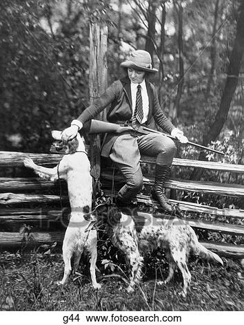 1920S Woman Sits On Wood Fence Holding Shotgun Over One Knee Petting One Of Two Dogs Hunting Hat Fashion View Large Photo Image Picture - 1920S Woman Sits On Wood Fence Holding Shotgun Over One Knee Petting One Of Two Dogs Hunting Hat Fashion . Fotosearch