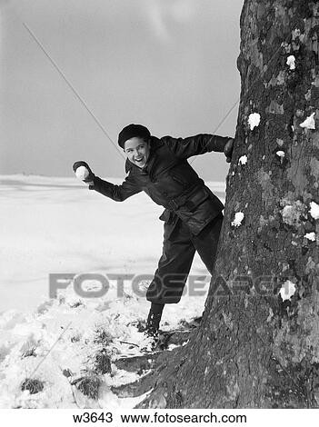 Stock Image - 1930 1930S Boy Looking Out From Behind Tree With Snowball In Hand Having Snowball Fight . Fotosearch