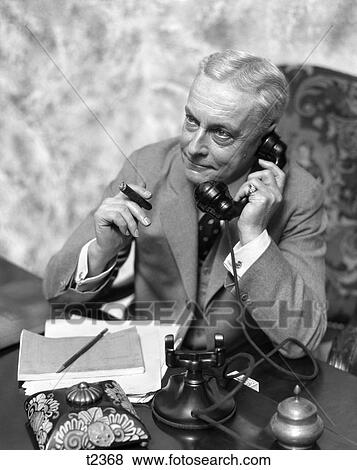 1930 1930S Elder Man Sitting At Desk Smoking Cigar Talking On Telephone View Large Photo Image Stock Photo - 1930 1930S Elder Man Sitting At Desk Smoking Cigar Talking On Telephone . Fotosearch